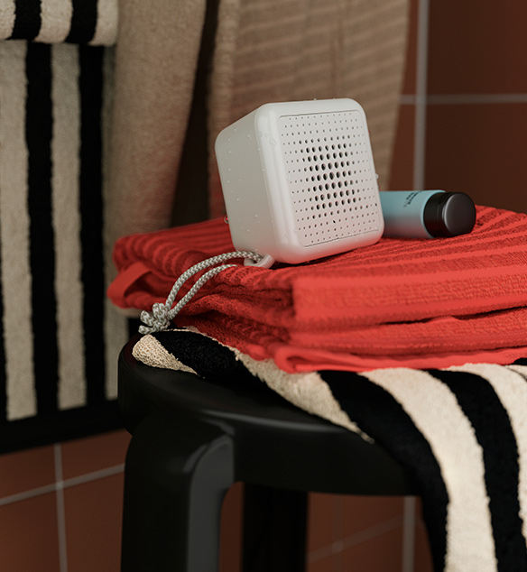 Un altavoz portátil Bluetooth VAPPEBY de color blanco, colocado sobre unas toallas dobladas de color óxido, encima de un taburete negro en un baño.