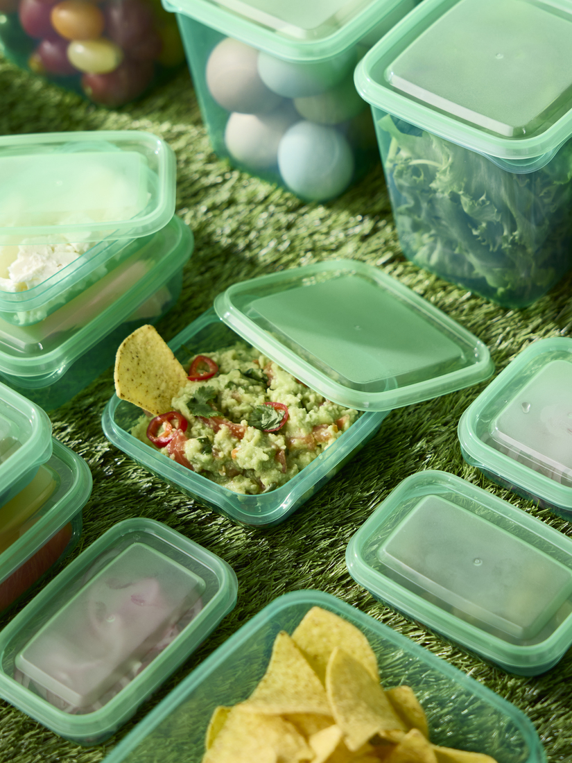 Top view of several green plastic food storage containers on artificial grass, with one containing guacamole and tortilla chips.