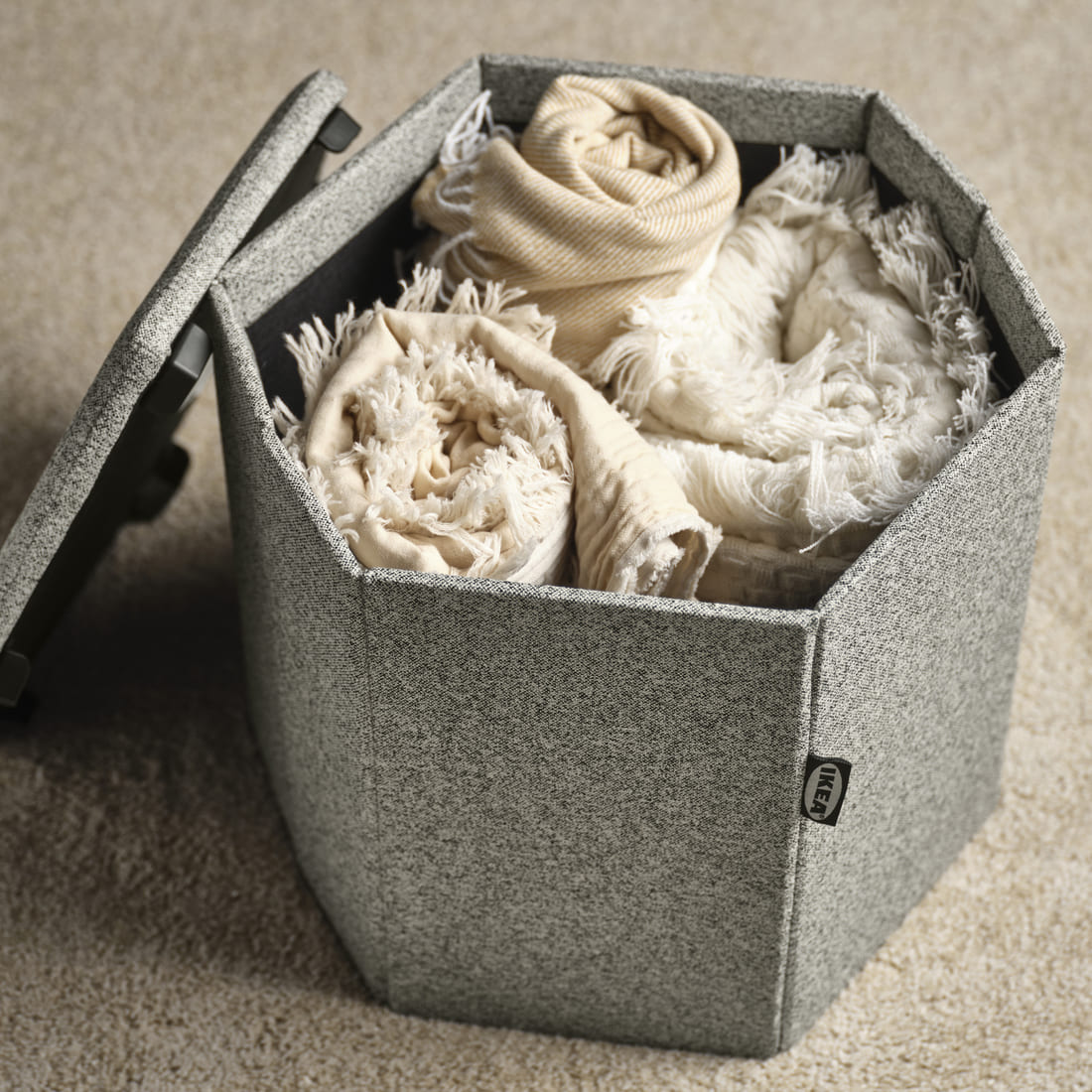 Hexagonal gray fabric storage box with its lid partially open, filled with rolled blankets and textiles on a beige carpet.