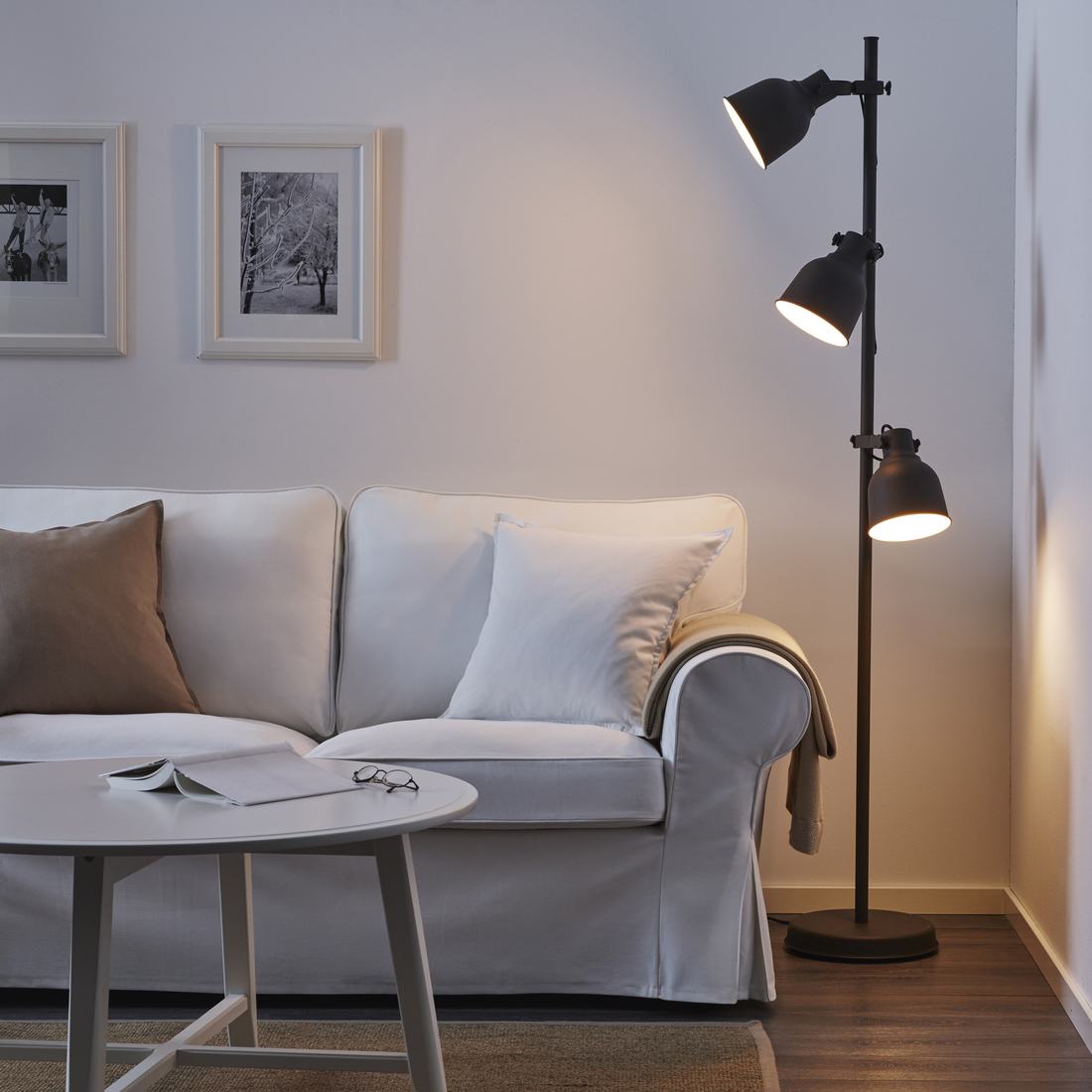 Black metal floor lamp with three adjustable spotlights in a cozy living room next to a white sofa and coffee table.