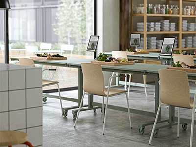 Mesas largas de color verde salvia con ruedas configuradas con sillas de madera, tablas de cortar con ingredientes y pantallas digitales, en un taller moderno y luminoso.