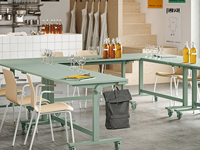 Mesas de color verde salvia con ruedas dispuestas en forma de U con sillas de madera, copas de vino y botellas, en un espacio de taller de cocina o sala de degustación.