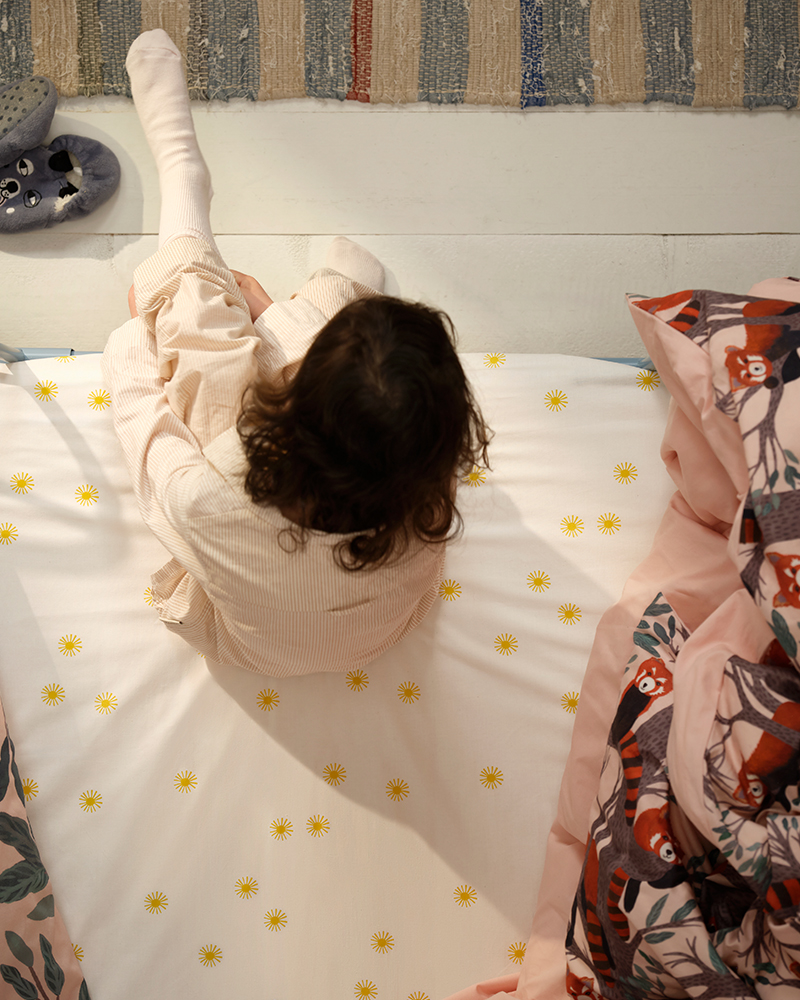 Una niña sentada en una cama con sábanas con motas amarillas, rodeada de cojines con dibujos de pandas rojos de la colección ALPTALL.