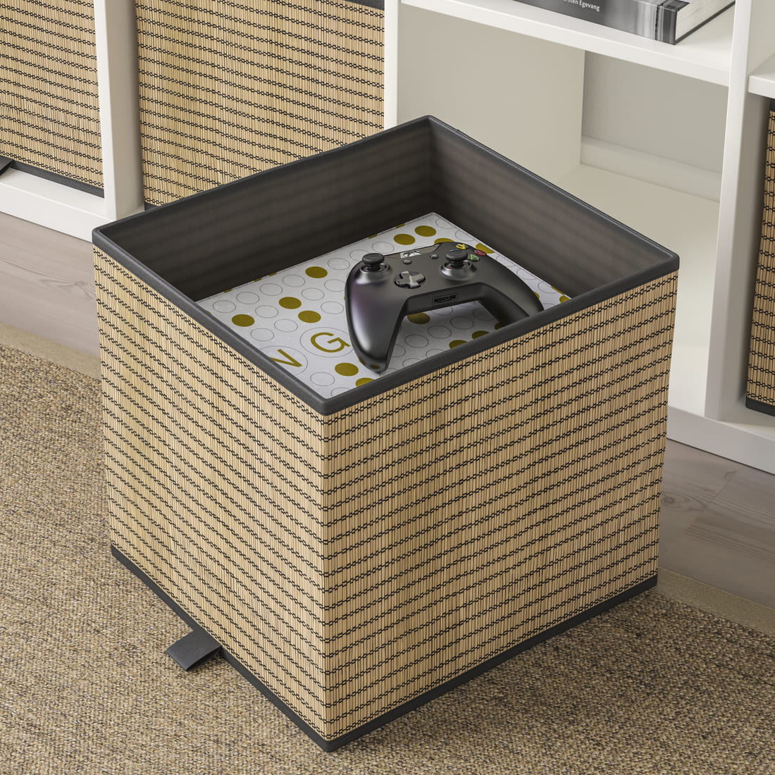 Caja de almacenaje cúbica de fibra natural tejida con bordes oscuros, con un mando de videojuegos en su interior junto a un mueble modular blanco.