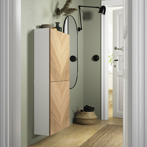Baño moderno con armario alto BESTÅ en madera clara, detalles en negro, cesta tejida en el suelo y planta frondosa.