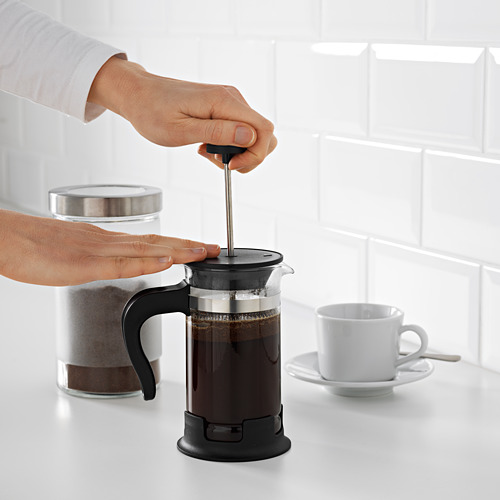 La persona presiona el émbolo en la cafetera de prensa francesa negra en el mostrador blanco; taza y bote cerca.