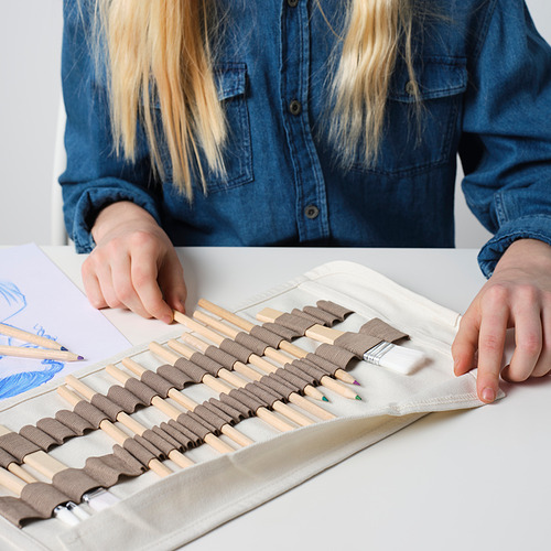 Una persona que viste una camisa azul usa un estuche de lápices para organizar los materiales de arte. El estuche, hecho de tela ligera, sostiene de forma segura lápices de colores y un pincel.