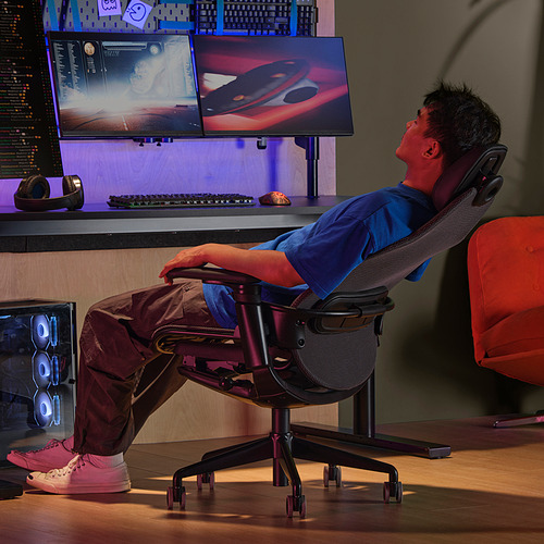 La persona se reclina en una silla de juego ergonómica con reposacabezas y respaldo de malla, frente a dos pantallas vibrantes.
