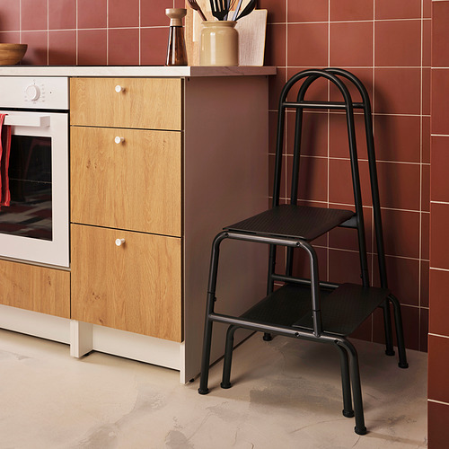 Escena de la cocina: pared moderna de azulejos marrones, mueble de madera clara con horno, taburete de 2 escalones de metal gris con escalones negros y pasamanos.