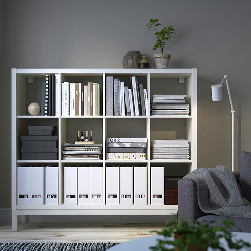 Estantería blanca KALLAX con artículos, entre sillón y lámpara, rematada con plantas.
