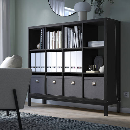 La estantería KALLAX negra contiene libros, carpetas y decoración. Es alto, rectangular, de líneas limpias. A su lado hay una silla gris.