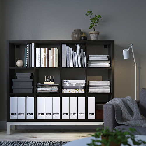 Una estantería negra KALLAX contiene libros, cajas y plantas; esquina de una habitación con un sofá gris y lámpara de pie.