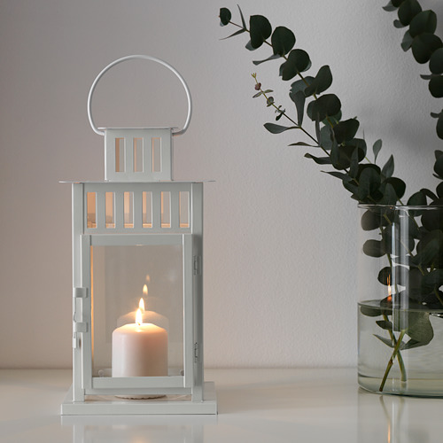 Linterna iluminada BORRBY sobre superficie blanca, cálido resplandor, diseño minimalista, eucalipto en jarrón transparente.