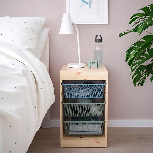 Mueble de madera TROFAST junto a la cama con lámpara, botella de agua y reloj. Tres cajones, edredón blanco sobre la cama.