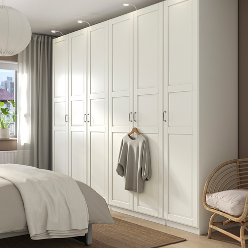 Un gran armario blanco PAX con cinco secciones contra una pared. Es un suéter colgante y se encuentra junto a una cama y una silla de mimbre.