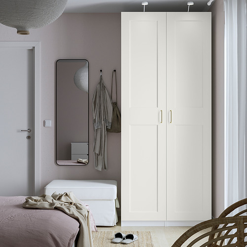 Dormitorio moderno con armario blanco GRIMO, espejo grande, ropa de cama ligera y silla de mimbre.