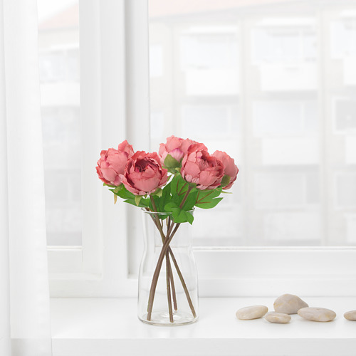 Jarrón transparente con flores artificiales rosas sobre una encimera blanca junto a una ventana.