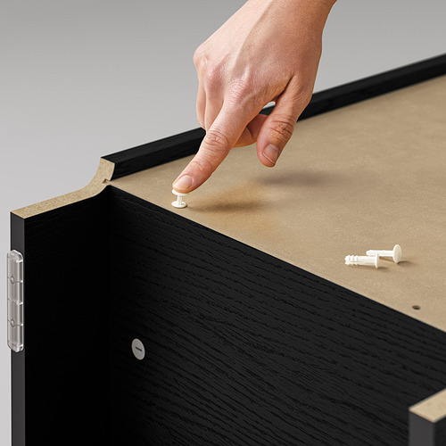 Colocando a mano una pequeña clavija blanca en una estantería negra de BILLY. La librería está hecha de aglomerado con acabado en madera.