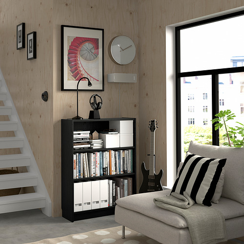 Acogedora habitación con librería negra BILLY, lámpara, auriculares, guitarra y reloj. Gran ventanal, cojín a rayas y luz natural brillante.