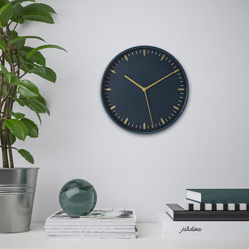 Reloj de pared con esfera azul oscuro y manecillas doradas, movimiento silencioso de cuarzo, sobre mesa con libros y plantas.