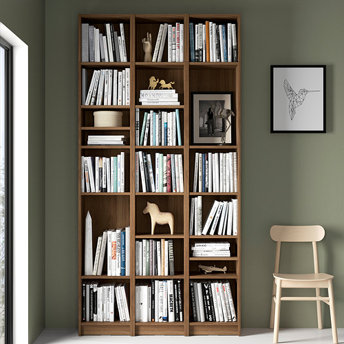 Gran estantería de madera BILLY llena de libros, decorada con pequeñas figuritas, contra la pared verde. Silla de madera suave al lado, arte de colibríes arriba.