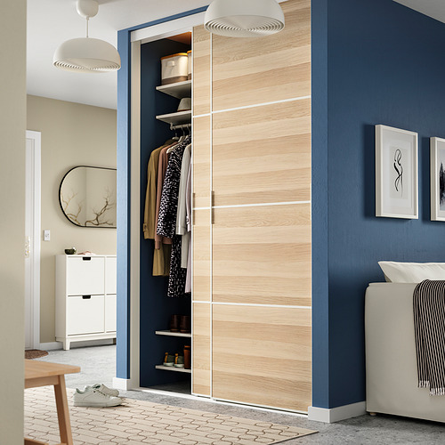 Armario de madera clara con puertas correderas, mostrando ropa y estanterías, junto a una silla y bajo lámparas colgantes.