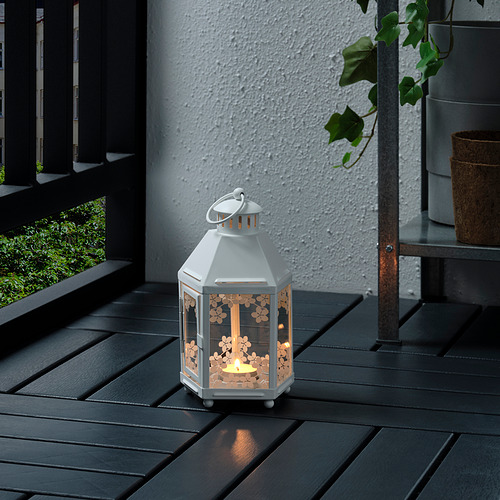 Una pequeña linterna blanca se encuentra sobre una mesa de madera oscura, proyectando una luz cálida a través de su patrón floral.