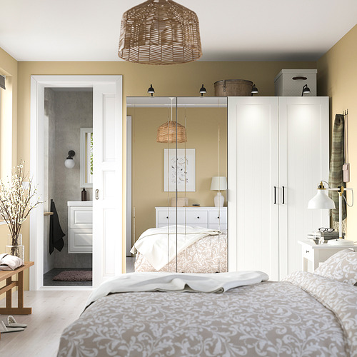Las puertas de espejo KAMMARTORP reflejan una cama hecha, una luz, una mesa, una alfombra gris y la puerta del baño en un dormitorio ordenado.