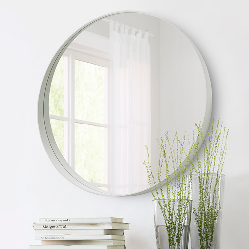 Un espejo redondo cuelga de una pared blanca reflejando una ventana. Debajo de él, se ve una pila de libros y un jarrón con plantas verdes.
