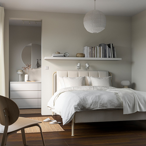 Dormitorio minimalista con ropa de cama blanca, estantería de libros, luz redonda, espejo y silla.