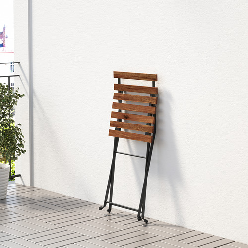 Silla plegable de madera con patas de metal negro apoyada en una pared blanca. Diseñado para uso en exteriores, resistente y fácil de almacenar.