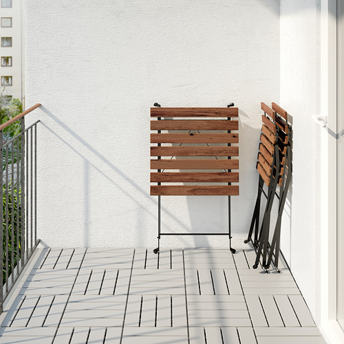 Montaje de balcón con mesa de madera TÄRNÖ plegada y sillas apiladas, apoyada en una pared blanca.