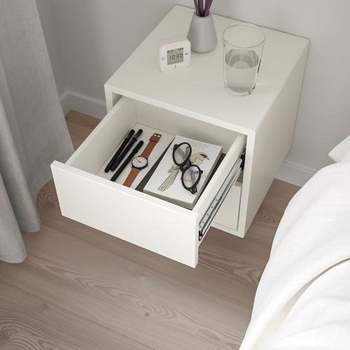 Mesita de noche blanca con cajón abierto. Arriba: reloj, vaso de agua, flores moradas. Cama y cortinas parcialmente vistas.