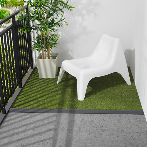 Pequeña silla de plástico blanco sobre alfombra de césped artificial verde cerca de barandilla negra.