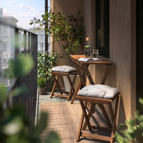 Pequeño balcón con mesa y taburetes plegables de madera marrón NÄMMARÖ, acolchados en blanco, colocados junto al follaje verde.