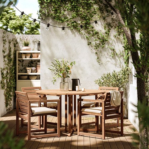 Juego de comedor al aire libre con mesa de madera y cuatro sillas acolchadas de color beige bajo guirnaldas de luces.
