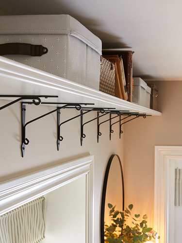 Estante de pared blanco con soportes de metal negro para guardar cajas de almacenamiento y archivos. Debajo, un espejo redondo refleja una planta verde en una habitación cálida.