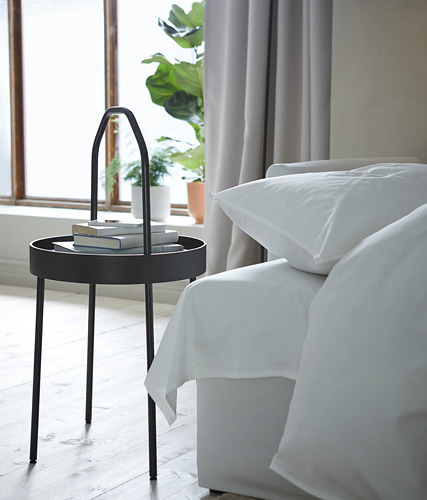 Mesita de noche BURVIK, negra, tapa de bandeja redonda, asa curvada, ropa de cama blanca, planta de interior junto a la ventana.