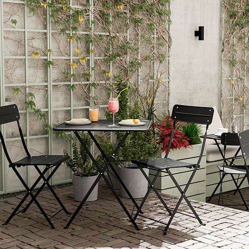 Silla de exterior SUNDSÖ negra con mesa, plegable, sobre un patio de ladrillo con plantas.