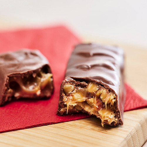 Dos barras de chocolate, una mordida, que revelan un caramelo crujiente y masticable y un relleno de nueces. Sobre una superficie roja.