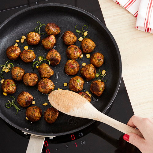Fríe las albóndigas veganas, dorándolas. Mezclas de espátula de madera; uñas rojas. Sartén negra, estufa controlada por el tacto.