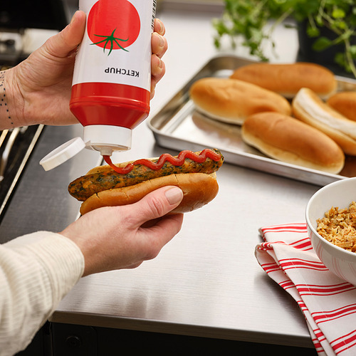Una persona exprime salsa de tomate roja en un perrito caliente que tiene en la mano.