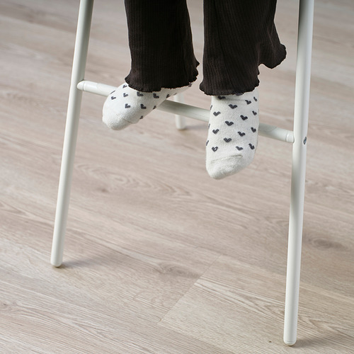 Silla junior blanca con los pies de alguien en calcetines estampados con corazones negros descansando sobre ella.