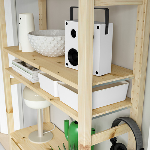 Unidad de almacenamiento de madera con estantes que sostienen un altavoz blanco, un tazón blanco, recipientes blancos, libros, regadera verde y lámpara blanca.