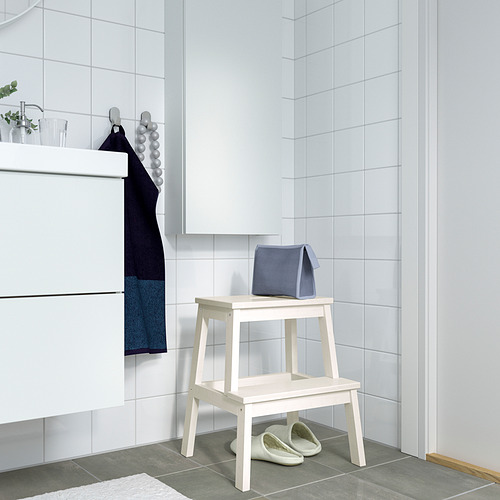 Pequeño taburete blanco junto a lavabo blanco, zapatillas grises, bolso azul y toallas.