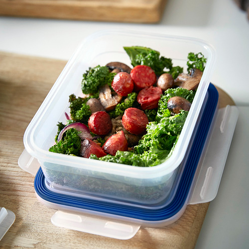 Un recipiente de plástico transparente para alimentos con tapa azul contiene una ensalada de col rizada, tomates, champiñones y salchichas, perfecta para almacenar las sobras.