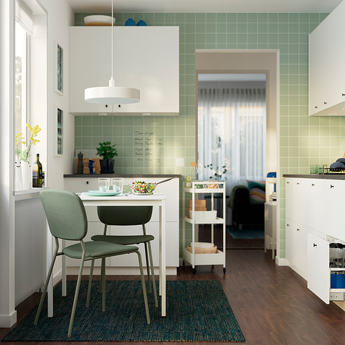 Una cocina moderna con una silla VIHALS verde. Cerca de una mesa con platos y un tazón, armarios blancos, azulejos verdes y suelos oscuros.