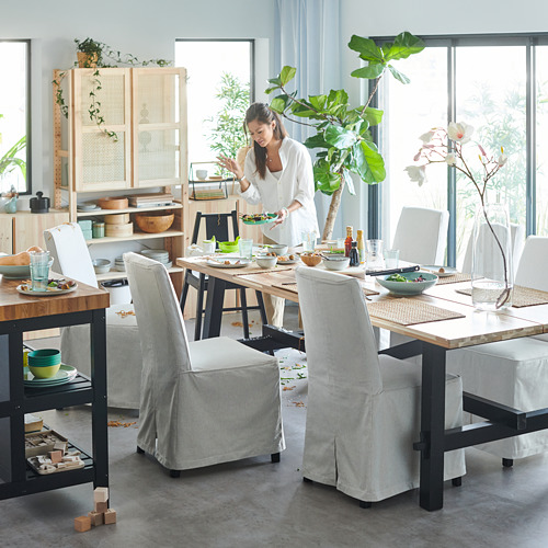 Mujer sirviendo comida en una cocina moderna con mesa SKOGSTA, sillas grises, estantes de madera y plantas verdes.