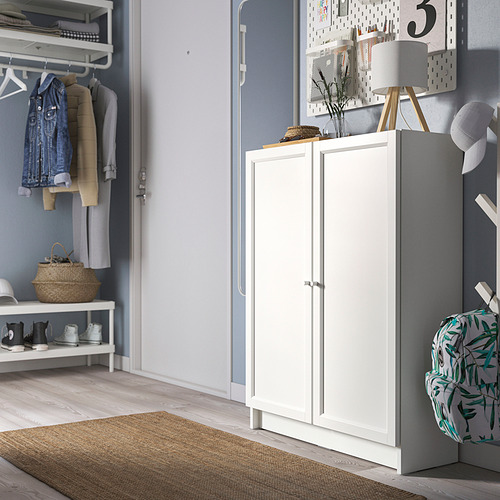 Librería blanca BILLY en entrada con puertas cerradas, junto a perchero, sobre alfombra beige, con cesta y mochila cerca, pared azul claro con tablero perforado.
