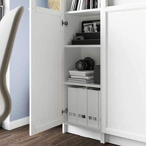 Estantería blanca BILLY con puerta de vidrio, que exhibe libros, una cámara y cajas de almacenamiento. Estantes ajustables a la vista, anclados a la pared.